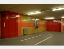 Unterirdischer Gang mit gelb-roten Fliesenwänden, roten Türen und Deckenleuchten. Eine Telefonzelle und ein Abfalleimer sind an der Wand befestigt, der Boden ist grau mit eingezeichneten Pfeilen.