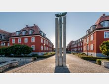 Ein gepflasterter Platz mit einem modernen Denkmal aus vertikalen Steinsäulen in der Mitte, umgeben von roten Wohnhäusern mit weißen Fensterrahmen und Satteldächern. Gepflegte Grünflächen und Laternen säumen den Platz.