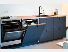 Moderne Küchenzeile mit dunklen, grifflochgefrästen Fronten und Holzarbeitsplatte, ausgestattet mit Einbaubackofen, schwarzer Spüle und hoher Armatur. Die Spülmaschine ist geöffnet und zeigt den Geschirrkorb; an der weißen Wand sind mehrere Steckdosen zu sehen.