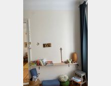 Heller Wohnraum mit schlichter weißer Wand, an der ein schmales Regal mit Büchern, Kerzen und Deko-Elementen befestigt ist. Darunter befinden sich ein geflochtener Korb, Meditationskissen und eine kleine Sitzbank; an der rechten Seite hängt ein dunkler Vorhang.