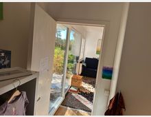 Ein Blick durch eine geöffnete Tür in ein hell erleuchtetes Wohnzimmer mit großem Fenster, das auf einen Garten blickt. Im Raum befinden sich ein dunkelblaues Sofa, ein Holzkasten, ein bunter Teppich und verschiedene Dekorationsgegenstände.