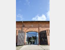 Ziegelsteinfassade eines Gebäudes mit einer großen Glastür in einem Rundbogen, flankiert von geöffneten dunklen Holztoren. Im Vordergrund stehen Dekorationselemente wie Pflanzen und Laternen, und ein Hund liegt auf dem Boden.