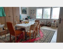 Esszimmer in einem Altbau mit einem rechteckigen Tisch, der mit einer Spitzentischdecke bedeckt ist, umgeben von sechs Stühlen. Der Raum ist mit einem großen Teppich, Holzschränken, alten Spielzeugmöbeln und Wanddekorationen ausgestattet; große Fenster bieten Blick auf ein Wohngebiet mit Bäumen und roten Dächern.