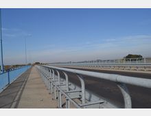 Eine breite Brücke mit blauem Geländer und Fahrbahn, ergänzt durch einen angrenzenden Gehweg. Der Himmel ist klar und blau, und im Hintergrund sind flache Landschaften und wenige Bäume sichtbar.
