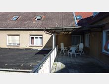 Eine gepflasterte Dachterrasse mit Vordach aus Metall und Glas, ausgestattet mit weißen Kunststoffstühlen und einem Tisch. Im Hintergrund sind ein beiges Gebäude mit Fenstern und ein Satteldach mit Dachfenstern zu sehen.