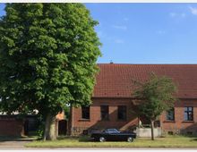 Backsteingebäude mit roten Dachziegeln in einer ländlichen Umgebung, eine große Kastanie und ein kleiner Baum im Vordergrund, daneben ein schwarzer Oldtimer auf einer Wiese geparkt.