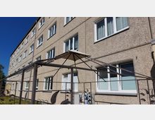 Mehrstöckiges Wohnhaus mit einer Fassade aus grauem Putz und mehreren Fenstern mit weißen Rahmen. Im Vordergrund steht ein Pavillon-Gerüst aus Metall mit einem spitzen Stoffdach sowie dekorativen Elementen.