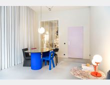 Minimalistisch eingerichteter Raum mit hellen Wänden, einem lilafarbenen Türrahmen und einem großen Fenster mit weißen Sprossen. Ein ovaler Tisch mit blauem Sockel und verschiedenen Stühlen steht mittig, daneben ein Beistelltisch mit einer roten Lampe und Büchern; links ist ein weißer Vorhang zu sehen.