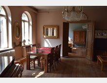 Esszimmer in einem historischen Gebäude mit Holzboden, einem Holztisch und Stühlen. Die Wände sind beige, dekoriert mit einem vergoldeten Spiegel und Rundbogenfenstern; eine offene Tür führt in einen zusätzlichen Raum mit einem großen Holzschrank.