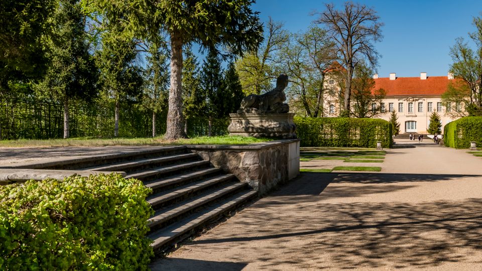 Formal garden with a wide gravel path bordered by trimmed hedges and tall trees, featuring stone steps and a statue on a raised terrace. In the background, a large historic palace-like building with a red tiled roof stands under a clear blue sky.