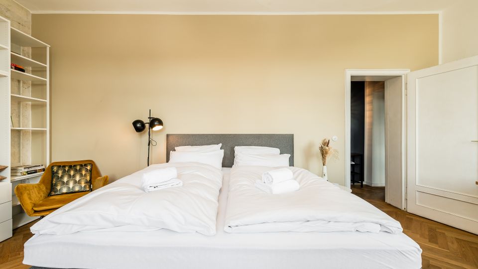 Helles Schlafzimmer mit zwei zusammengestellten Einzelbetten mit weißer Bettwäsche und grauem Kopfteil. Links stehen ein offenes Regal, ein gepolsterter Sessel und eine schwarze Stehlampe; rechts führt eine offene Tür in einen Flur, der Boden ist aus Holzparkett.