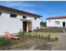 Zwei einfache, weiße Wohngebäude mit Flachdach und roten Dachziegeln, die in einem offenen Bereich mit sandigem Boden stehen. Zwischen den Gebäuden befinden sich ein kleiner Garten mit Blumen, ein Liegestuhl und ein kleiner Tisch mit Stühlen.