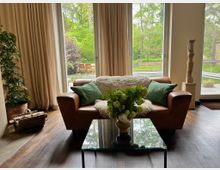 Helles Wohnzimmer mit bodentiefen Fenstern und beigen Vorhängen, davor ein braunes Ledersofa mit grünen Kissen und einer weißen Decke sowie ein Glastisch mit Blumen. Durch die Fenster ist eine Terrasse und ein grüner Garten mit Bäumen zu sehen; daneben stehen eine Zimmerpflanze, ein Korb mit Holzscheiten und eine Skulptur auf einem Sockel.