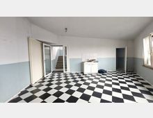 Ein leerer Innenraum mit schwarz-weiß kariertem Fliesenboden, hellgrauen Wänden und einer kleinen weißen Spüle mit Ablage in einer Ecke. Rechts sind ein Fenster und eine offene Tür, links eine Glastür, die zu einer Treppe führt.