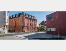 Straßenszene mit einem orangefarbenen, mehrstöckigen Altbau mit Mansardendach auf der linken Seite und einem modernen Gebäude mit roter Klinkerfassade und blauer Aufschrift "Union" rechts. Im Vordergrund befinden sich Straßenlaternen, ein Verkehrsschild und gepflasterte Gehwege.