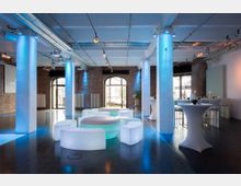 Moderner Veranstaltungsraum mit gemauerten Wänden, großen Rundbögenfenstern und einer offenen Decke mit sichtbaren Rohren. Der Raum ist mit LED-Beleuchtung in Blau und Weiß akzentuiert und verfügt über gebogene Sitzmöbel mit Beleuchtung sowie hohe Stehtische.