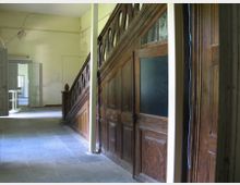 Ein Flur in einem alten Gebäude mit einer hölzernen Treppe auf der rechten Seite und verglasten Holztüren darunter. Der Boden besteht aus Steinfliesen, und im Hintergrund sind offene Doppeltüren sichtbar, durch die Tageslicht einfällt.