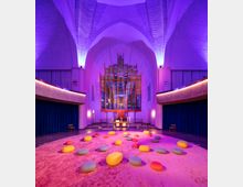Innenraum einer modern beleuchteten Kirche mit hoher gewölbter Decke, violettem Licht und einem zentralen bunten Glasfenster hinter dem Altar. Auf einem pinken Teppich liegen bunte, runde Sitzkissen verteilt, und blau-goldene Vorhänge bedecken seitlich die Wände.