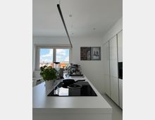 Moderne Küche mit einer weißen Arbeitsplatte und einem eingebauten Herd mit schwarzer Glaskeramikoberfläche. Im Hintergrund befinden sich ein großes Fenster, Küchengeräte wie ein Mixer und eine Kaffeemaschine sowie weiße Schränke an der rechten Wand.