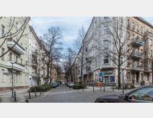 Straßenszene in einem städtischen Wohngebiet mit mehrstöckigen Altbauhäusern, einigen mit Balkonen. Im Vordergrund sind kahle Bäume, parkende Autos und ein Schild mit der Aufschrift "Helmstraße" zu sehen.