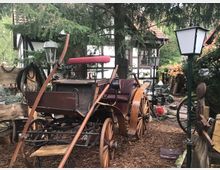 Historische Kutsche mit Holzrädern und rotem Sitz in einem rustikalen Hof mit Kopfsteinpflaster, Laternen und Fachwerkgebäude im Hintergrund, umgeben von Bäumen und Dekorationsgegenständen.