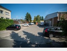 Gepflasterter Parkplatz in einem Gewerbegebiet, umgeben von einem Wohngebäude mit bewachsener Fassade auf der linken Seite und einer Lagerhalle mit Wellblechdach auf der rechten Seite. Mehrere parkende Autos, Bäume im Hintergrund und Container stapeln sich am hinteren Rand des Geländes.