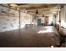 Großer, heller Raum mit industriellem Charme, gekennzeichnet durch weiße Ziegelwände mit verblasster Beschriftung, eine Holzfußbodenoptik und freiliegende Deckenbalken. Der Raum ist spärlich möbliert mit einem Holztisch und Stühlen und enthält industrielle Details wie Metalllampen, Heizungsrohre und eine historische Waage an der Wand.