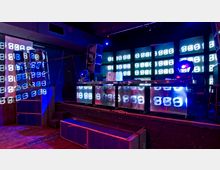 Elektronischer Clubraum mit einer Bühne, auf der ein DJ-Pult steht. Die Wände und der Tisch sind mit leuchtenden Displays ausgestattet, die abstrakte Lichtmuster zeigen, während die Szenerie in blau-violettem Licht beleuchtet wird.