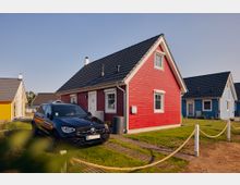 Ein rotes, einstöckiges Holzhaus mit Satteldach steht in einem Wohngebiet, umgeben von anderen farbigen Häusern. Ein schwarzer PKW ist vor dem Haus auf einer kleinen Einfahrt geparkt, und der Rasen des Grundstücks ist mit einem Seilgeländer abgegrenzt.
