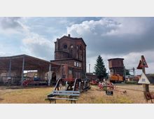 Industrielles Gelände mit historischen Backsteingebäuden, darunter ein mehrstöckiger Bau mit Rundfenstern und ein Wasserturm. Im Vordergrund alte Bahnausrüstung und Schilder, die auf den Eisenbahnbezug hinweisen.