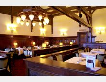 Gemütlicher Speiseraum mit dunkler Holztäfelung, weißen Tischdecken und gepolsterten Stühlen. An der Decke hängt ein großer Kronleuchter, und wandmontierte Lampen sorgen für warmes Licht; eine Tür mit der Aufschrift „Toiletten“ ist im Hintergrund sichtbar.