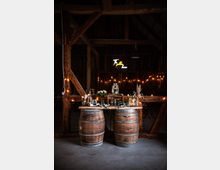 Rustikale Innenansicht einer Scheune mit Holzbalken, zwei Holzfässern als Basis für einen dekorierten Tisch mit Blumen, Getränkespendern und Kerzen. Im Hintergrund leuchten Lichterketten und ein Neon-Schild mit der Aufschrift "The golden hour".