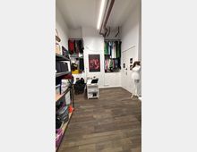 Small costume storage room with white walls and dark wood flooring, featuring shelves of bins on the left and two recessed wall closets with hanging clothes at the back. Overhead exposed pipes and a long fluorescent light run along the ceiling, with a poster on the back wall and a mannequin stand near a set of white doors on the right.