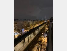 Nächtliche Stadtansicht mit schneebedeckten Straßen und Gebäuden, einer erhöhten U-Bahn-Trasse in der Mitte und dem Berliner Fernsehturm im Hintergrund. Straßenlaternen und Fenster erleuchten die Szene.
