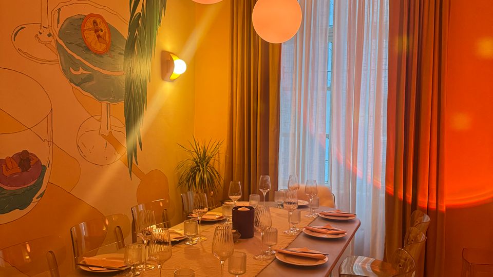 Warm-lit private dining room with a long table set for multiple guests, clear acrylic chairs, and two round pendant lights overhead. A large wall mural decorates one side, and tall curtains cover a floor-to-ceiling window at the end of the room.