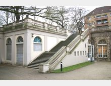 Gebäude mit klassizistischer Architektur, hell verputzt, mit einer breiten Außentreppe, verzierten Balustraden und halbkreisförmigen Fenstern. Im Vordergrund ein Gehweg und eine kleine Rasenfläche, im Hintergrund Bäume und ein mehrstöckiges Wohngebäude.