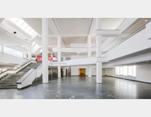 Weitläufiger, moderner Innenraum mit hohen weißen Decken, breiten Treppen aus dunklem Stein und großen Fenstern, die viel Tageslicht hereinlassen. Die Architektur ist von offenen Galerien und klaren geometrischen Linien geprägt.