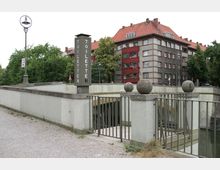 Öffentlicher Toiletteneingang mit Betonmauern, Metallgeländern und runden Betonkugeln als Dekoration; im Hintergrund Wohnhäuser mit roten Dachziegeln und Bäumen.