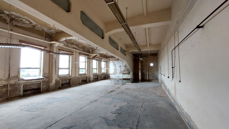 Verlassener, leerer Raum in einem Industriebau mit abblätternder Farbe an Wänden und Decke, mehreren großen Fenstern entlang einer Wand, sichtbaren Kabelrohren und abgewittertem Betonboden.