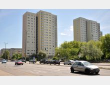 Zwei hohe Wohngebäude mit gelb-beiger Fassade und Balkonen stehen an einer breiten Straße, die von Autos befahren wird. Im Vordergrund befinden sich eine Ampelkreuzung und Grünflächen mit Bäumen.