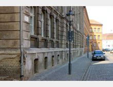 Historische Straßenszene mit einem langen, mehrstöckigen Gebäude aus verwittertem Stein, kleinen Kellerfenstern und dekorativen Fensterrahmen. Auf dem Kopfsteinpflaster stehen Laternen, ein geparktes Auto und Wegweisschilder, im Hintergrund ein rotes Backsteingebäude.