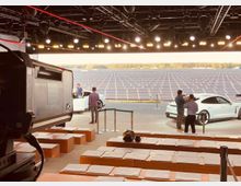 Aufnahme eines Präsentationsbereichs mit Sitzreihen im Vordergrund, einer Bühne mit zwei weißen Autos und Personen im Mittelgrund, und einem großen Feld mit Solarpanels im Hintergrund. Die Szenerie ist unter einem mit Scheinwerfern ausgestatteten Studiodach beleuchtet.