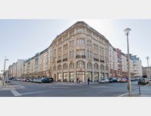 Eckgebäude in einer Stadt mit einer historischen Steinfassade und großen, bogenförmigen Fenstern im Erdgeschoss. Links und rechts befinden sich angrenzende, mehrstöckige Wohn- und Geschäftsgebäude; vor dem Gebäude stehen Autos entlang der Straßenseite.