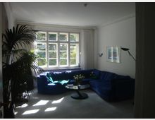 Wohnzimmer mit einer großen, blauen Eckcouch, einem runden Glastisch mit Blumen darauf und einem großen Fenster mit weißen Vorhängen, das viel Tageslicht hineinlässt. Links steht eine Zimmerpflanze, und an der rechten Wand hängen Bilder sowie eine Lampe.