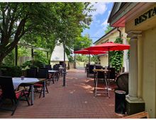 Außenbereich eines Restaurants mit roten Sonnenschirmen, schwarzen Tischen und Stühlen auf einer roten Pflasterfläche. Im Hintergrund sind Bäume, eine helle Gartenmauer, ein Kamin und ein Pergoladach zu sehen, die die gemütliche Atmosphäre unterstreichen.