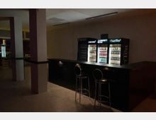 Dunkler Innenraum einer Bar oder Lounge mit langem Tresen und zwei hohen Barhockern. Hinter dem Tresen stehen beleuchtete Getränkekühlschränke, daneben sind Fenster und breite Säulen im Raum zu sehen.