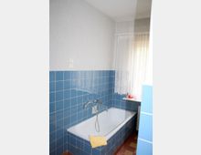 Badezimmer mit einer weißen Badewanne, umgeben von hellblauen Fliesen. An der Wand befindet sich ein Wasserhahn mit Handbrause, und am Fenster hängt eine weiße Spitzen-Gardine, die das Tageslicht leicht dämpft.
