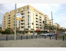 Mehrstöckiges Wohn- und Geschäftsgebäude in heller Farbe mit Balkonen und großen Fenstern an einer Kreuzung. Im Erdgeschoss befinden sich Geschäfte und ein Restaurant, davor führen Straßen mit parkenden und fahrenden Autos sowie Gehwege mit einigen Fahrrädern entlang.