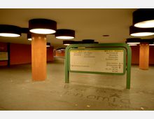Ein unterirdischer Fußgängertunnel mit orange gefliesten Säulen und rechteckigen Deckenleuchten. Im Vordergrund steht ein grünes Wegweisschild mit Informationen zu Ausgängen und öffentlichen Verkehrsmitteln.