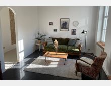 Wohnzimmer mit einer antiken grünen Samtcouch, einem kleinen Holztisch auf einem weißen Teppich und einem roten Sessel mit floralem Muster. An der weißen Wand hängen Bilder und eine Wanduhr, daneben steht eine Stehlampe. Ein großes Fenster lässt Tageslicht in den Raum, und ein seitlicher Durchgang führt zu einem weiteren Raum mit einer Mauer aus Sichtziegeln.